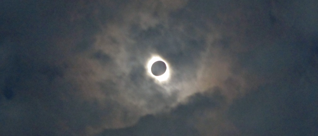 Solar eclipse during totality. Black ball with cloudy white corona around it in an otherwise darkened cloudy sky.