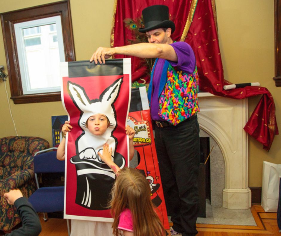 Magician Owen at a birthday party magic show holding his bunny banner while a child poses using it.