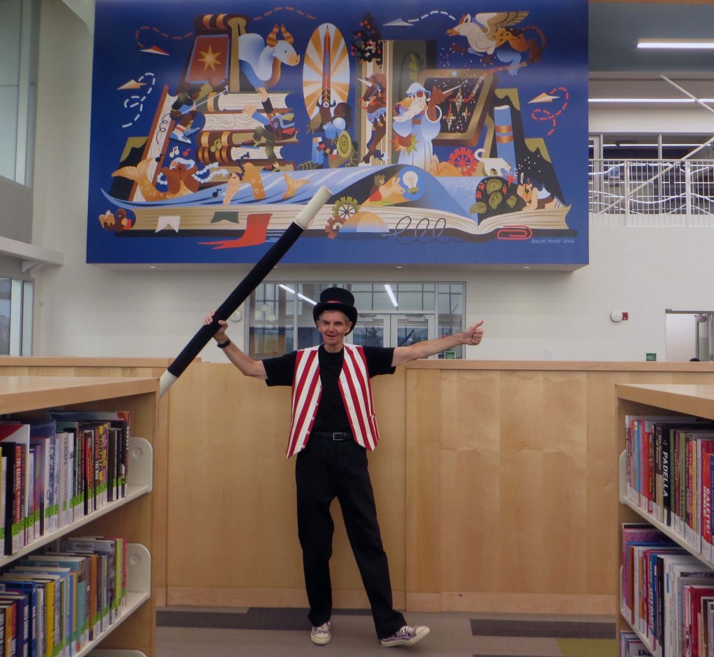 Owen posing in front of large library theme wall mural at Oakville Public Library's newly opened Sixteen Mile branch.